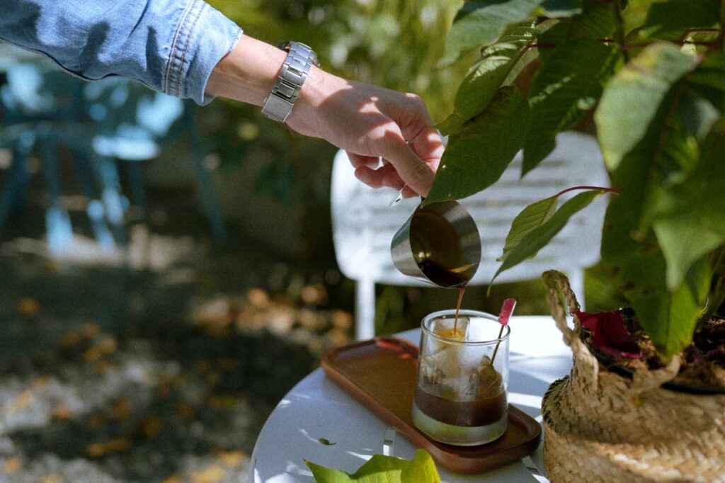 A person pours coffee into a glass in a garden setting, surrounded by lush greenery.